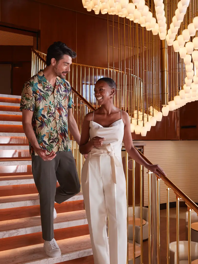 Two guests walking down a sweeping brass-and-marble staircase beneath a cascade of warm lantern lights